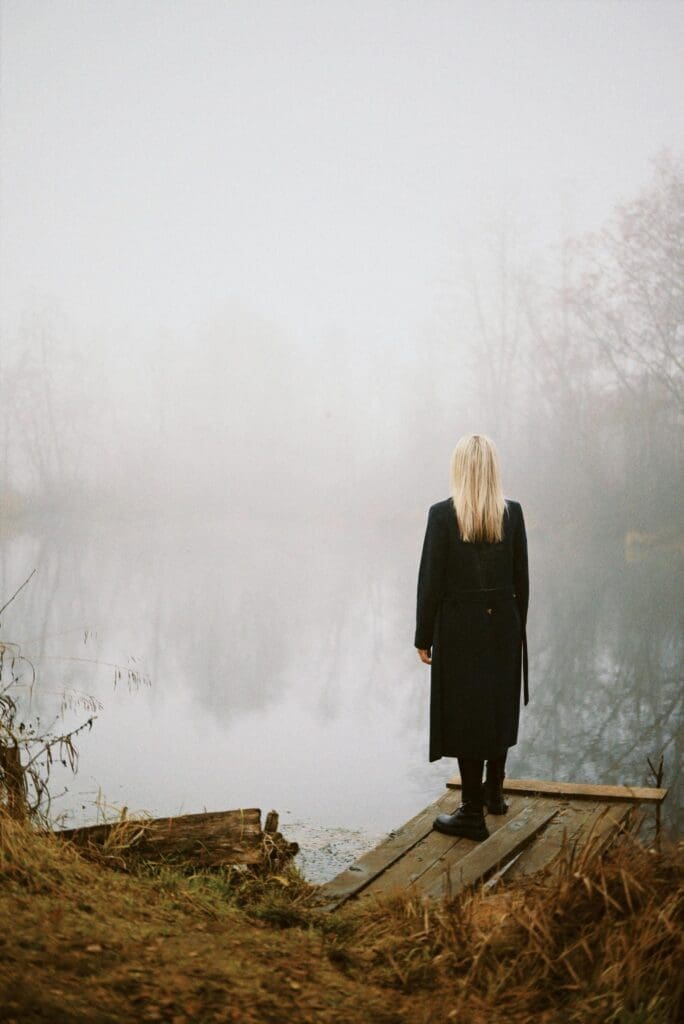 Blonde woman standing on a dock overlooking foggy lake, minimal and calm atmosphere. Scandinavian-inspired setting. 